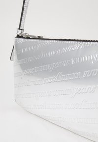 Sølv patentlædertaske med præget "tommyjeans" logo, rektangulær form, lynlåslukning og en kort skulderrem.