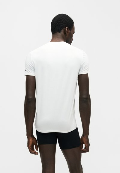 Άνδρας με κοντά σγουρά μαλλιά που φοράει μια απλή λευκή κοντομάνικη μπλούζα και μαύρα σορτς, στέκεται με την πλάτη προς τα εμπρός σε λευκό φόντο.