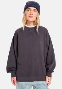 Donkergrijze oversized sweatshirt met ribgebreide manchetten, ronde hals en een klein logo detail. Met patroon bedekte lichtblauwe geruite broek.