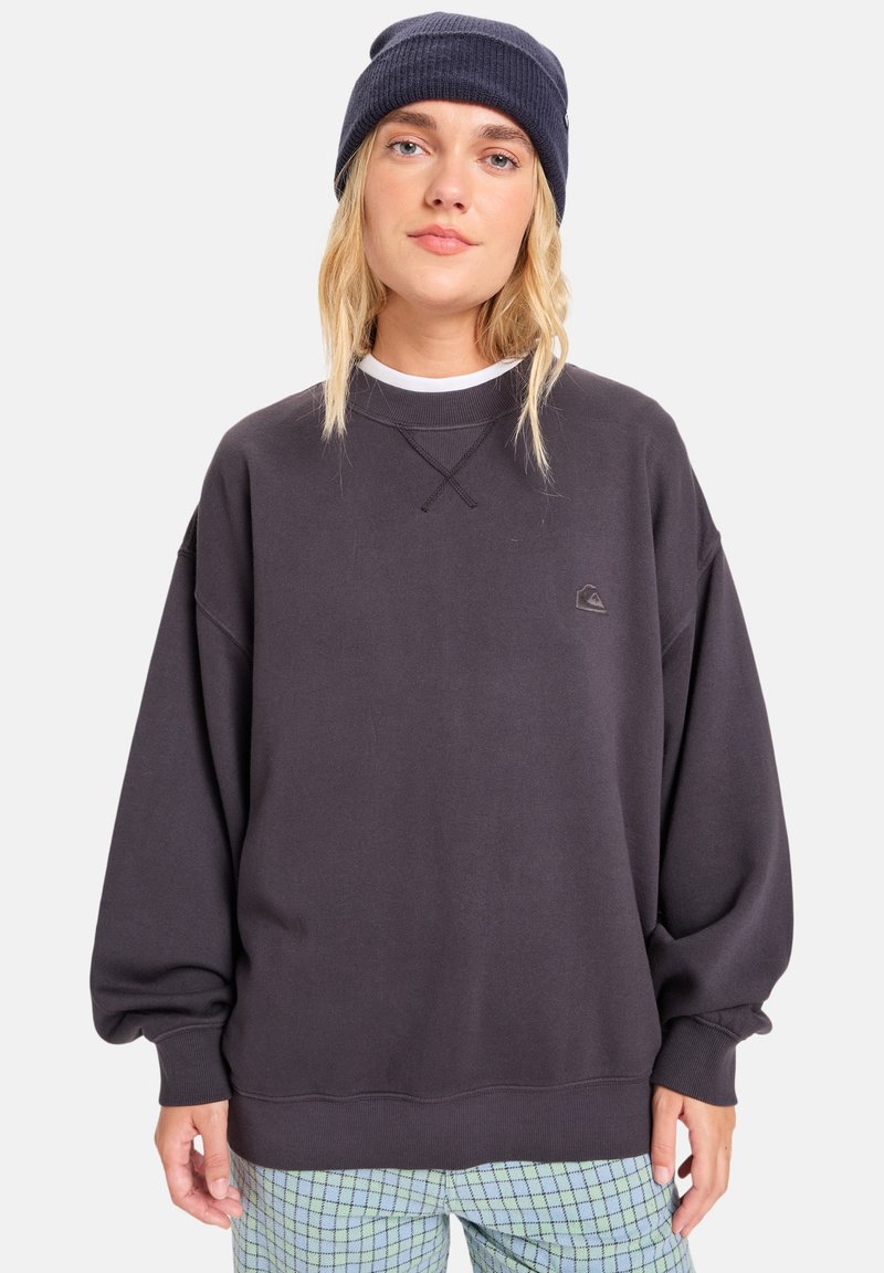 Donkergrijze oversized sweatshirt met ribgebreide manchetten, ronde hals en een klein logo detail. Met patroon bedekte lichtblauwe geruite broek.