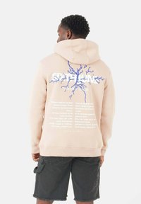 Felpa leggera rosa chiaro con design grafico blu e testo sul retro. Il materiale è morbido, con una vestibilità comoda e tasca a marsupio frontale.