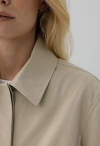 Beżowy płaszcz z kołnierzem o gładkiej, matowej fakturze z czystymi szwami i minimalistycznym wykończeniem, pokazany od szyi w górę.