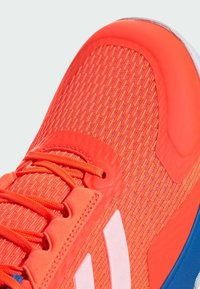 Scarpa atletica di un vivace arancione con parte superiore testurizzata, strisce bianche a contrasto e una sovrapposizione liscia sulla punta con accenti blu.