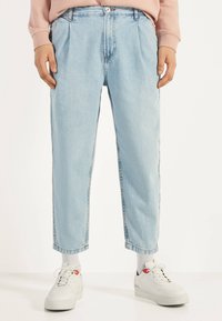 Vaqueros de mezclilla azul claro con un ajuste relajado, frente plisado y longitud recortada, combinados con zapatillas blancas y calcetines grises.