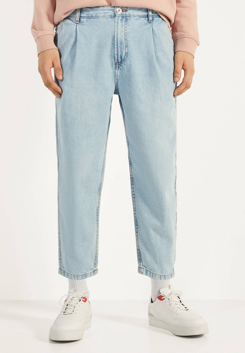 Vaqueros de mezclilla azul claro con un ajuste relajado, frente plisado y longitud recortada, combinados con zapatillas blancas y calcetines grises.