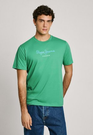 T-shirt verde in cotone con maniche corte e scollatura a girocollo, caratterizzato dal logo "Pepe Jeans London" in azzurro. Indossato con jeans in denim blu.