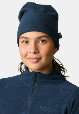 Fleece hue i marineblå med blød tekstur, let slouchy design og et logo-tag på siden, bæres med en matchende fleecejakke.