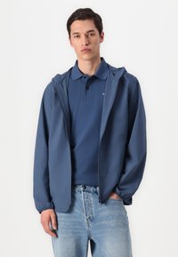 Jachetă ușoară de culoare bleumarin cu glugă, fermoar frontal, manșete elastice, purtată peste un tricou polo asortat, asociată cu blugi de culoare albastră deschisă.