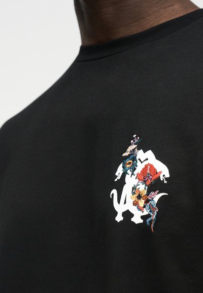 Μαύρο πουκάμισο με λευκά στυλιζαρισμένα γράμματα και πολύχρωμα κεντημένα λουλούδια στο στήθος, που φοριέται από άτομο με σκουρόχρωμο δέρμα.