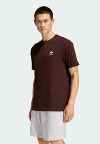 Hnedočervené bavlnené tričko s okrúhlym výstrihom a krátkymi rukávmi, na hrudi s bielym logom Adidas. Nosené s svetlosivými šortkami.