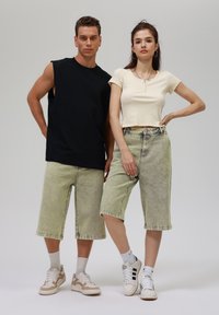 Two models wearing casual outfits. Male in a black sleeveless shirt, light green denim shorts; female in a light yellow cropped top, green shorts.