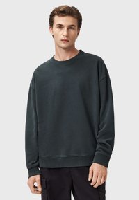 Felpa verde scuro realizzata in morbido tessuto, con scollo a girocollo, spalle scese e maniche e orlo a coste. Il modello indossa pantaloni neri.