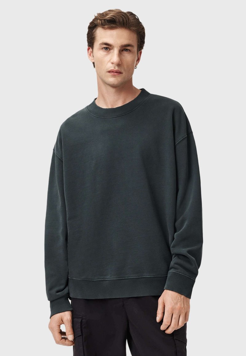 Felpa verde scuro realizzata in morbido tessuto, con scollo a girocollo, spalle scese e maniche e orlo a coste. Il modello indossa pantaloni neri.