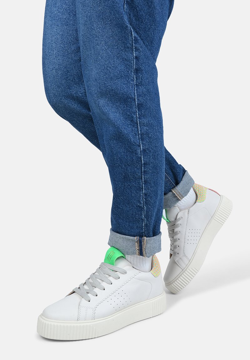 Sneakers bianche con una finitura testurizzata, dettaglio verde sulla linguetta e motivo multicolore sul tallone; abbinate a jeans blu arrotolati.