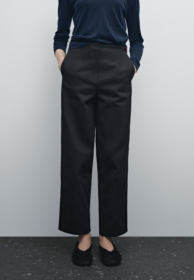 Personne portant un pantalon noir à jambes larges, des chaussures noires pelucheuses sans lacets et un haut sombre à manches longues, debout contre un mur gris uni.