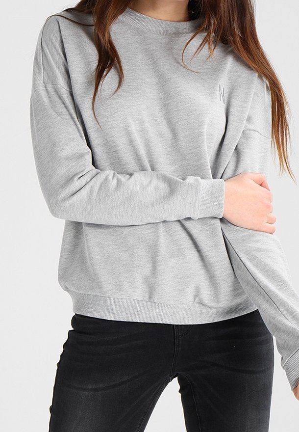 Personne portant un sweat-shirt gris clair à manches longues et un pantalon noir, tenant son bras gauche avec la main droite sur le torse.