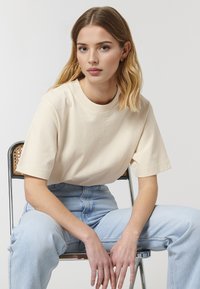 Beige, kortærmet, bomulds t-shirt med rund hals, iført lyseblå højtaljede jeans, siddende på en metalstol.