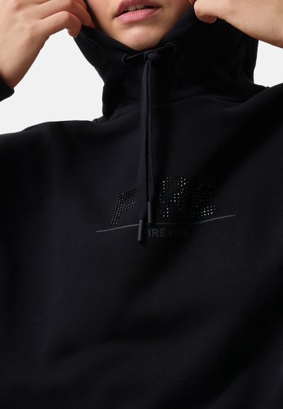 Person passt einen schwarzen Hoodie an, auf dem sich das "FIRE+ICE"-Logo aus kleinen schwarzen Strasssteinen auf der Brust befindet, vor weißem Hintergrund.