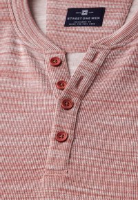 Chemise henley à rayures rouges et blanches avec une texture côtelée, dotée d'une patte de boutonnage avec quatre boutons rouges et d'une étiquette de marque tissée.