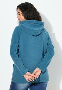 Donna con capelli ricci che indossa un hoodie blu, in piedi di profilo con le mani unite dietro la schiena.