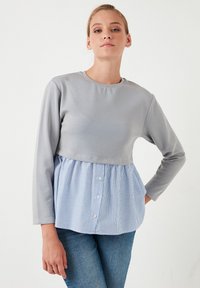 Sivi top s dugim rukavima i naboranim donjim dijelom u plavo-bijelim prugama. Ima okrugli izrez i detalj s gumbima sprijeda.