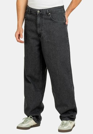 Locker sitzende schwarze Jeans aus Denim, getragen mit hellen Sneakers, Frontansicht von der Taille bis zu den Füßen vor hellem Hintergrund.