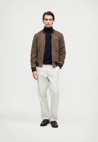 Young man standing with hands in pockets wearing brown suede bomber jacket, navy turtleneck, white trousers, and black shoes.