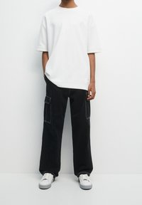 Witte oversized t-shirt met korte mouwen, gecombineerd met zwarte cargobroek met zakken en wit stiksel, en witte sneakers.