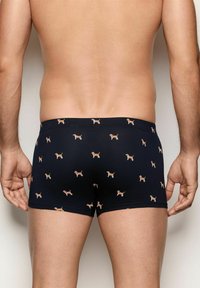 Rückansicht eines Mannes in marineblauen Boxershorts mit kleinem braunem Hundemuster, der vor einer schlichten hellen Wand steht.