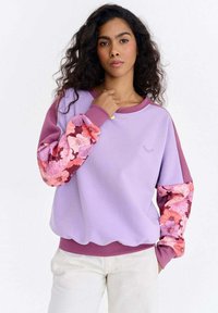 Lilla dressipluus, mille varrukad on roosade ja burgundia lillemustritega. Omaduseks on ümar kaelus ning soonikkäiste ja -äärega varrukad ja ääris. Sile kangapind.