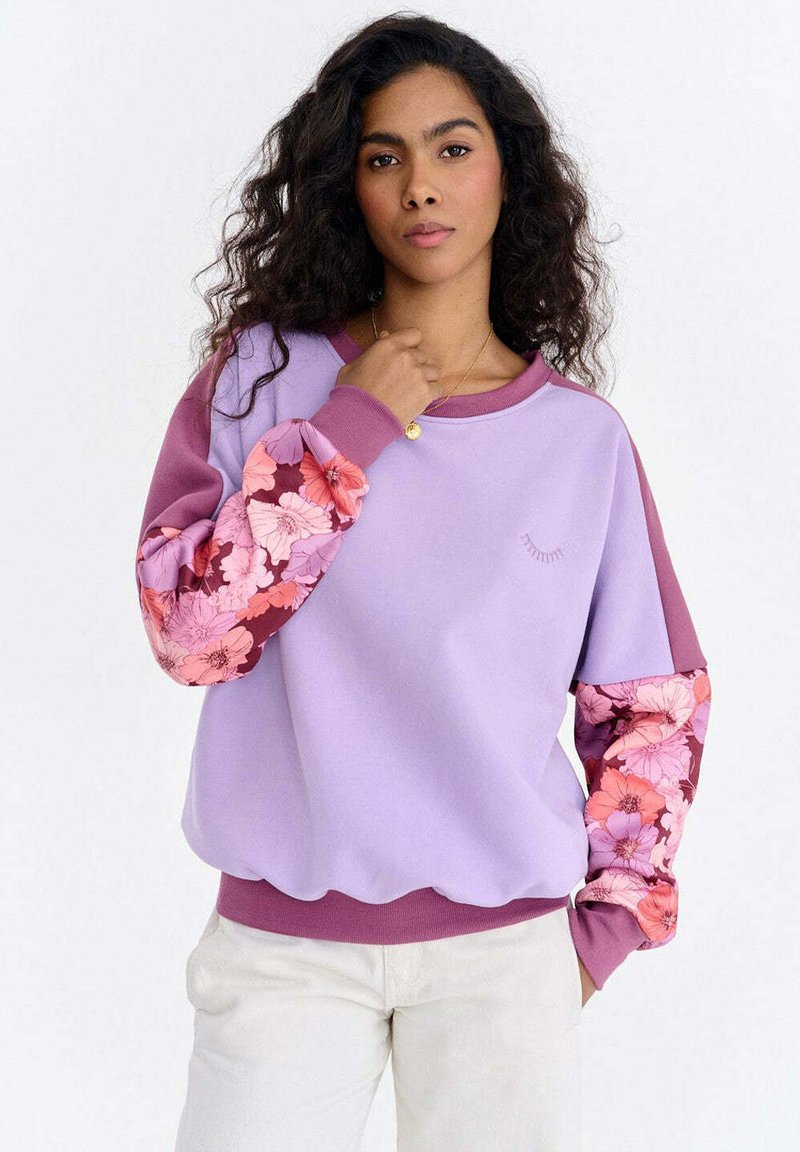 Lilla dressipluus, mille varrukad on roosade ja burgundia lillemustritega. Omaduseks on ümar kaelus ning soonikkäiste ja -äärega varrukad ja ääris. Sile kangapind.