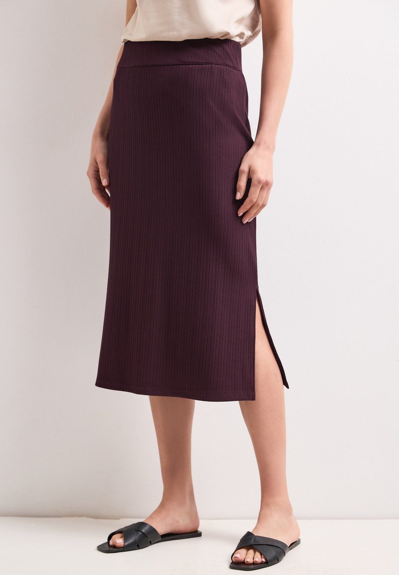 Femme portant une jupe midi côtelée violet foncé avec une fente latérale, un haut beige et des sandales plates noires, debout contre un mur uni.