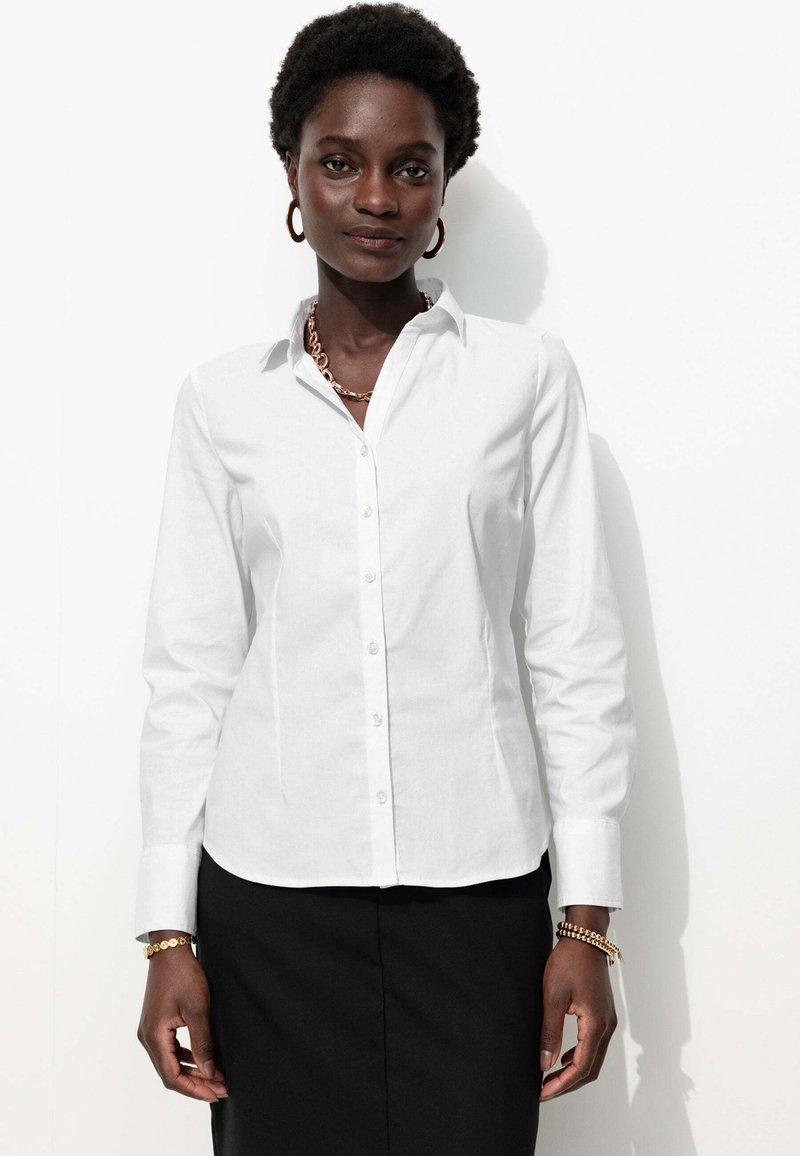 Chemise blanche à manches longues et à boutons, confectionnée dans un tissu lisse, avec un col pointu et une coupe subtile, associée à une jupe noire.