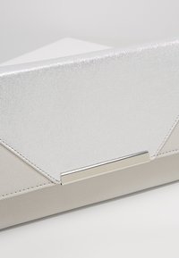 Kabelka typu clutch s texturovaným stříbrným vrchem, hladkým béžovým spodkem, angularním designem a štíhlým kovovým akcentem.