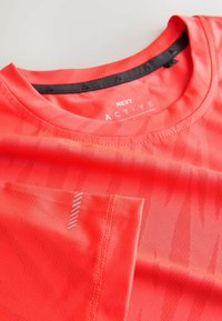 T-shirt de sport corail vif avec motif subtil, encolure ronde, manches courtes et petit détail réfléchissant sur la manche, étiqueté « NEXT ACTIVE ».