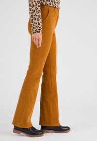 Pantalons en velours côtelé marron évasés avec une texture lisse et une taille haute, associés à des chaussures noires avec une semelle épaisse.