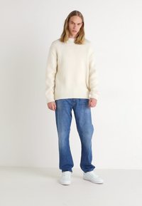Crème ribgebreide trui gecombineerd met lichtblauwe spijkerbroek en witte sneakers. De trui heeft een ronde hals en een ontspannen pasvorm.
