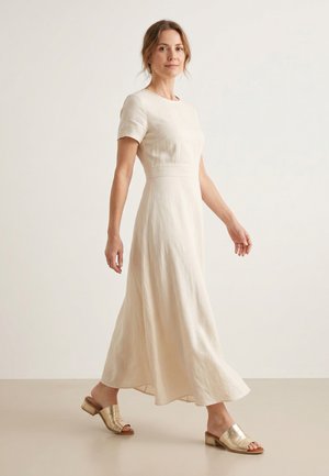 Kvinde iført en lyse beige midi-kjole med korte ærmer og guldfarvede lave, åbne mule-sandaler mod en ensfarvet lys baggrund.