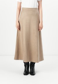 Femme portant une longue jupe beige, un haut blanc et des bottes noires mi-mollet, se tenant devant un fond blanc uni.
