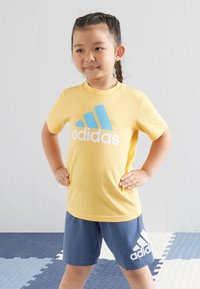 Keltainen puuvillapaita, jossa on sininen Adidas-logo; yhdistetty tummansinisiin shortseihin, joissa on valkoinen Adidas-kuvio. Lapsi seisoo kädet lanteilla.
