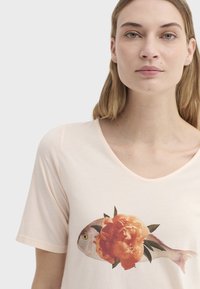 T-shirt à manches courtes de couleur rose pâle, présentant un graphisme de poisson avec une fleur orange et des feuilles vertes à l'avant. Texture douce en coton.
