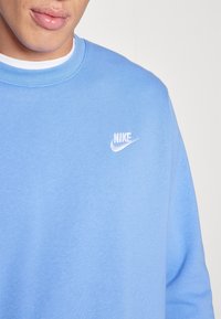 Sudadera de algodón azul claro con cuello y puños acanalados. Presenta un logo blanco de Nike en el lado izquierdo del pecho. Textura suave, diseño de cuello redondo.