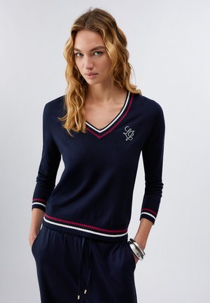 Młoda kobieta o falowanych blond włosach, ubrana w granatowy sweter z dekoltem w serek, ozdobiony czerwono-białymi paskami i monogramem, z rękami w kieszeniach, na jednolitym tle.