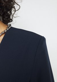 Blazer marine avec une conception d'épaules structurées et une texture lisse, associé à un collier en chaîne argenté, court jusqu'au cou.