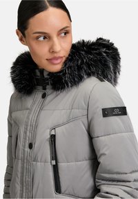 Doudoune grise rembourrée avec une capuche en fausse fourrure noire, dotée de poches zippées et d'accents de marque. Texture lisse et design matelassé.