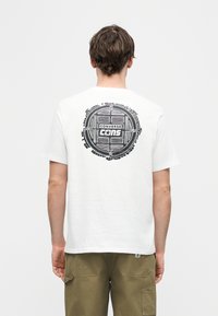 Άνδρας με λευκό T-shirt που φέρει μαύρο κυκλικό λογότυπο "Converse Cons" στην πλάτη, φοράει χακί cargo παντελόνι, στέκεται μπροστά σε απλό λευκό φόντο.