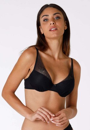 Donna con capelli lisci castani che indossa un reggiseno nero con dettagli in pizzo, posando con le mani giunte contro uno sfondo chiaro e uniforme.