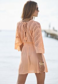 Licht perzikkleurige jurk met een kanten rug, korte fladdermouwen en een striksluiting. De stof heeft een gekreukelde textuur, wat de flow versterkt.