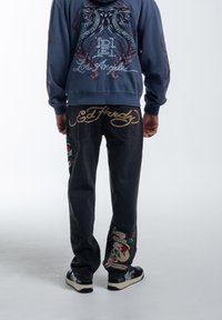 Donkerdenim jeans met geborduurde schedel en bloemenontwerpen, gecombineerd met een blauwe hoodie met een dubbele slanggrafiek en logo.