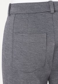 Šedé kalhoty z látky s vzorem a texturovaným povrchem, mají zadní kapsu a upravený pás. Detailní záběr na tkaninu.